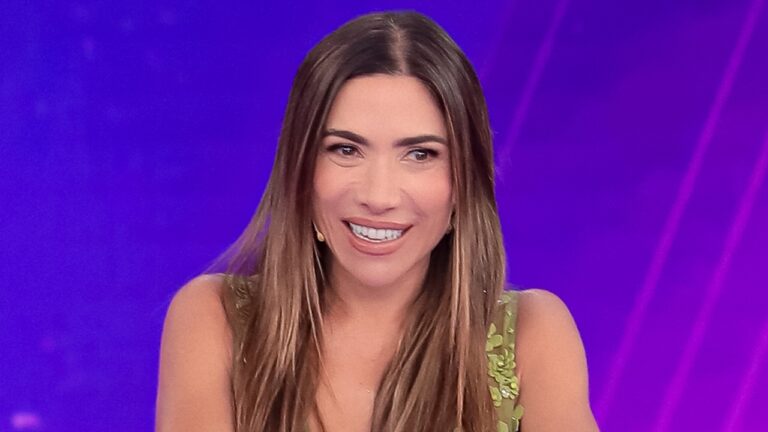 Patricia Abravanel, uma mulher sorridente com cabelos longos castanhos, aparece no estúdio do Programa Silvio Santos com fundo roxo iluminado, usando vestido verde e microfone headset.