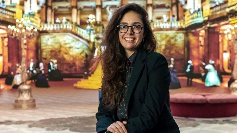 Uma mulher está sentada e sorrindo para a câmera, usando óculos e roupas formais escuras, como blazer. Ela tem cabelos longos e ondulados. Ao fundo, há um cenário que lembra um grande salão clássico ou palácio, com escadarias, iluminação quente e várias pessoas ao longe, algumas vestidas de forma elegante, sugerindo um ambiente histórico ou teatral.