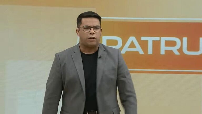 Apresentador de óculos e blazer cinza fala em estúdio de telejornal com painel laranja ao fundo exibindo programa Balanço Geral
