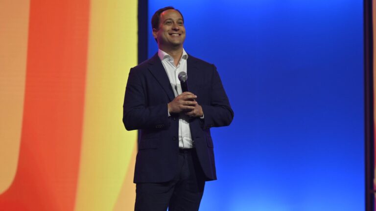 Paulo Marinho, diretor-presidente da Globo, veste terno e segura microfone, sorrindo em palco com fundo colorido em degradê durante evento do conglomerado.