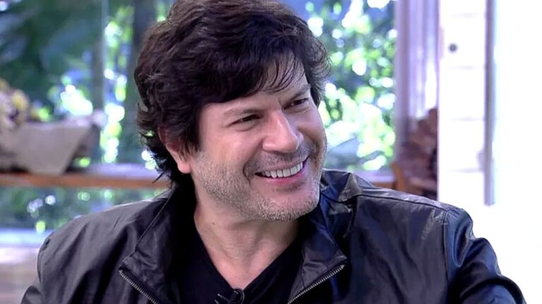 Homem sorri durante entrevista em programa de televisão com cenário iluminado e área verde ao fundo