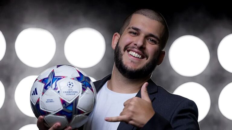 Homem sorrindo, com barba curta e cabelo raspado, segurando uma bola de futebol (da Liga dos Campeões) e fazendo um gesto de “hang loose” com a mão, diante de um fundo com luzes circulares fortes.