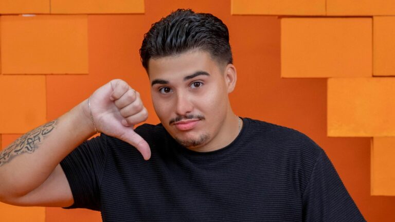 Pedro Henrique com camiseta preta faz gesto de desaprovação com polegar para baixo diante de fundo laranja em estúdio