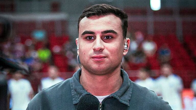 Um homem aparece em primeiro plano durante uma transmissão, olhando diretamente para a câmera. Ele usa um fone de ouvido intra-auricular e segura um microfone, indicando que está trabalhando como repórter ou comentarista. Veste uma blusa de moletom escura. Ao fundo, há uma arquibancada com várias pessoas desfocadas, sugerindo um ambiente esportivo ou ginásio.