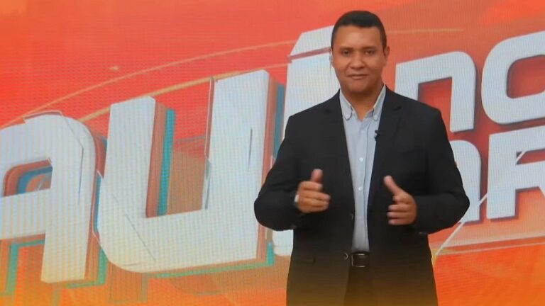 Apresentador gesticula em estúdio com telão laranja ao fundo exibindo nome de programa policial ao vivo