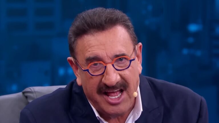 Ratinho, um homem de meia-idade com bigode e óculos coloridos, fala com expressão enfática no estúdio do SBT, usando terno escuro e microfone de lapela, com fundo azul desfocado.
