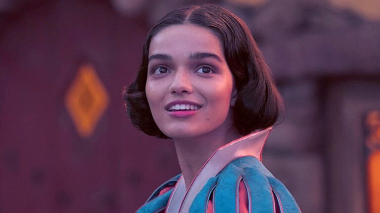 Mulher jovem de cabelos curtos e escuros sorri com expressão encantada enquanto olha para algo fora de quadro, usando figurino colorido de estilo clássico; ao fundo, cenário desfocado com iluminação suave.