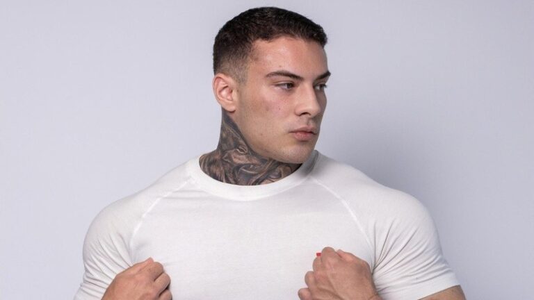 Rafa Marttinz posa de perfil em fundo branco, usando camiseta clara e exibindo tatuagens detalhadas no pescoço, com mãos segurando a gola da peça