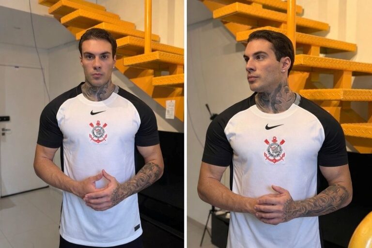 Rafa Marttinz veste camisa branca do Corinthians com mangas pretas, exibe tatuagens no pescoço e braços, posicionado à frente de uma escada amarela clara