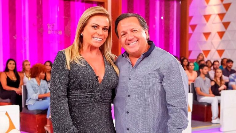 Apresentadores sorrindo abraçados em programa de auditório com plateia ao fundo e cenário colorido