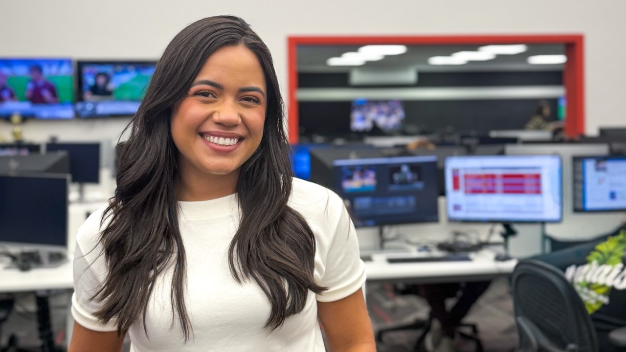 Jornalista Raisa Martins sorri de camiseta branca na redação da ESPN, com monitores de edição, computadores, bancadas de trabalho e TVs ao fundo ao fundo