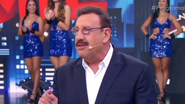 Apresentador Ratinho de terno azul marinho e óculos coloridos fala ao microfone no palco do SBT, com bailarinas de vestidos azuis ao fundo do cenário
