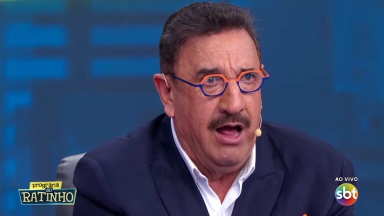 Ratinho, apresentador de televisão com bigode e óculos de armação colorida, fala durante programa ao vivo no estúdio. Ele usa terno escuro e microfone de lapela. No canto da tela aparece o logotipo Programa do Ratinho e o selo ao vivo do SBT.