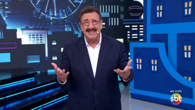 Ratinho, usando terno escuro e óculos redondos, fala ao público com as mãos abertas em gesto expressivo. Ele está em um estúdio com cenário estilizado de cidade à noite ao fundo, iluminado em tons de azul, com a indicação de transmissão ao vivo no canto da tela.