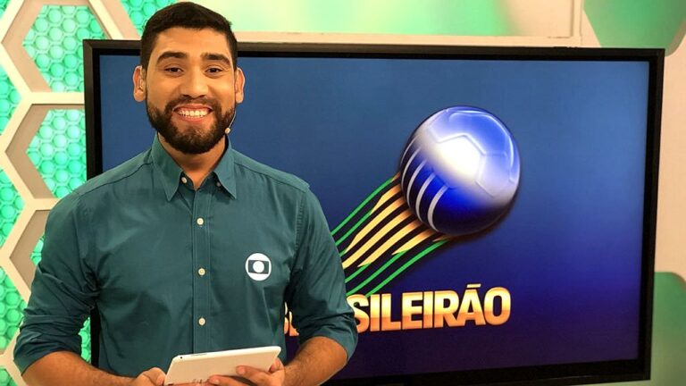 Apresentador esportivo sorri com tablet na mão diante de tela com logo do Brasileirão em estúdio de TV