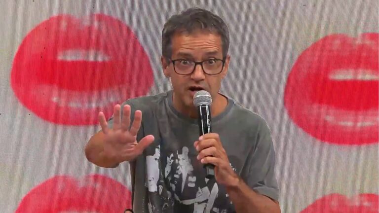 Ricardo Mendonça, de óculos e camiseta cinza, segura um microfone enquanto fala, com uma das mãos levantada em gesto de pare. Ao fundo, há um painel claro com grandes figuras vermelhas em formato de boca, parte do estúdio do Perrengue do Dia.