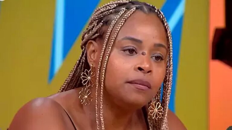 Uma mulher aparece em close em um estúdio de televisão, com expressão séria e atenta. Ela tem cabelo trançado com mechas claras e usa brincos grandes em formato de flor. Veste uma roupa de alça fina, deixando os ombros à mostra. O fundo é colorido, com tons vibrantes e formas geométricas, típicos de programas de entretenimento. A iluminação é forte e uniforme, destacando seu rosto e detalhes dos acessórios.