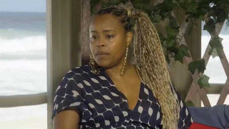 Mulher com tranças longas e brincos grandes olha para o lado com expressão séria enquanto está sentada em ambiente interno próximo a uma janela com vista para o mar