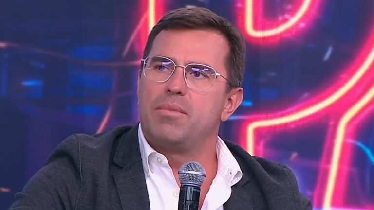 Rodrigo Bocardi encara a lateral com expressão séria, usa óculos de grau com armação transparente e veste blazer cinza sobre camisa branca em frente a um microfone