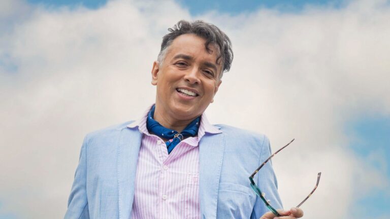 Ator Rodrigo Fagundes como Gigi na novela Volta por Cima, sorri usando blazer azul claro, camisa listrada rosa e lenço azul, sob um céu com nuvens