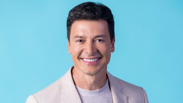 Rodrigo Faro sorri em retrato com fundo azul usando blazer claro e camiseta neutra em foto promocional