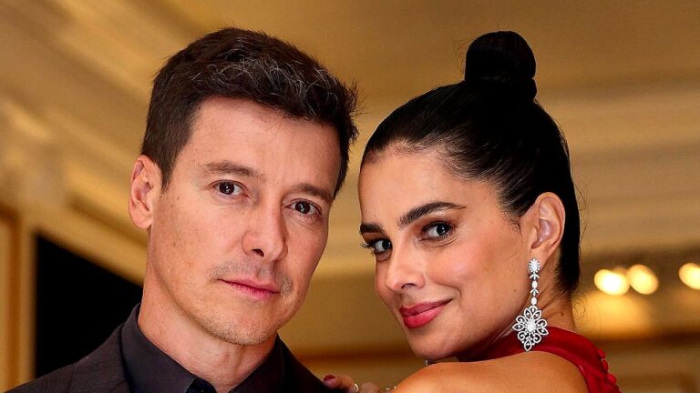 Homem e mulher posam juntos para foto em ambiente interno; ele usa terno escuro e expressão séria, enquanto ela, com cabelo preso em coque e vestido vermelho, sorri levemente usando brincos longos.