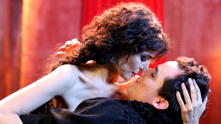 Casal se aproxima para beijo em cena romântica ambientada em salão com cortinas vermelhas ao fundo