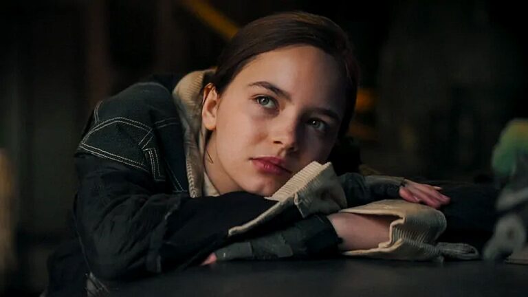 Uma jovem está apoiada sobre uma mesa, com a cabeça descansando sobre os braços cruzados. Ela usa um casaco escuro com capuz e uma blusa clara por baixo. Seu olhar está voltado para o lado, com expressão pensativa ou contemplativa. A iluminação é suave e o ambiente ao fundo é escuro e desfocado, sugerindo um espaço interno e silencioso, possivelmente um quarto ou sala pouco iluminada.