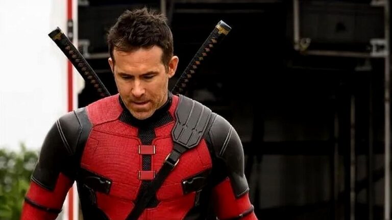 Homem com traje vermelho e preto de super-herói caminha com expressão séria, com duas espadas cruzadas presas nas costas em cena de filme de ação