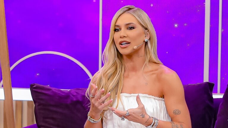 Virginia Fonseca, uma mulher loira com vestido claro e microfone, fala na última edição do programa Sabadou com Viginia, sentada em sofá com cenário roxo ao fundo.