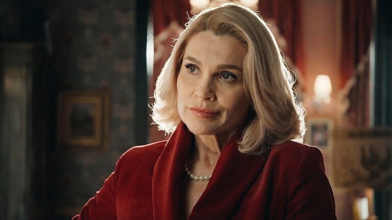 Mulher loira de expressão séria usa blazer vermelho e colar de pérolas enquanto conversa em ambiente interno sofisticado, com iluminação quente e decoração elegante ao fundo.