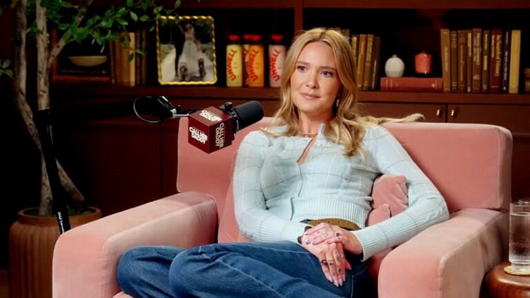 Mulher loira sentada em poltrona participa de entrevista em podcast com microfone à frente e estante de livros ao fundo