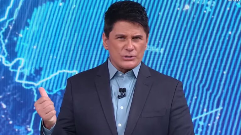 Cesar Filho, apresentador do SBT Brasil, veste um terno escuro e camisa azul. Ele fala em estúdio diante de um telão com mapa estilizado em tons de azul. Ele gesticula com a mão enquanto olha para a câmera durante a apresentação.