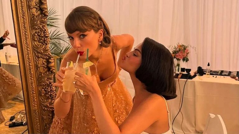 Duas mulheres elegantes compartilham bebida com canudos enquanto posam próximas a um espelho em ambiente interno decorado com flores e mesa de maquiagem ao fundo