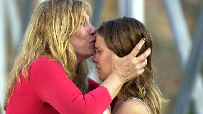 Mulher beija a testa de jovem emocionada enquanto segura seu rosto em gesto de carinho ao ar livre