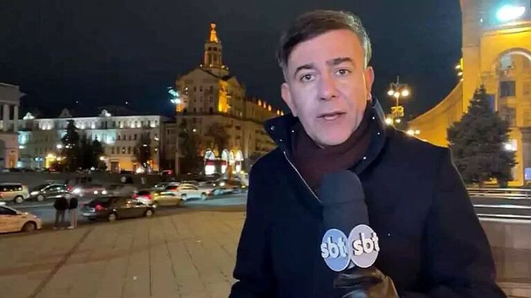 Texto Alternativo disse: Repórter do SBT fala ao vivo em praça europeia à noite com prédios iluminados e trânsito ao fundo