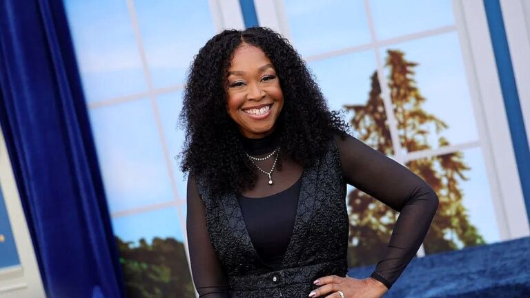 Uma mulher com cabelos longos e cacheados sorri enquanto está em um estúdio de televisão. Ela usa uma blusa preta de mangas transparentes com um colete escuro por cima, além de colares delicados. Está com uma das mãos apoiada na cintura, em uma postura confiante e descontraída. Ao fundo, há um cenário que imita grandes janelas com vista para árvores e céu, além de cortinas azuis, sugerindo um ambiente de programa de entretenimento ou talk show.