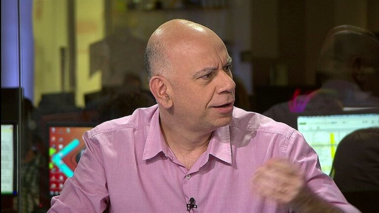 Homem careca veste camisa rosa e conversa em estúdio de televisão ou redação, com computadores e monitores ao fundo enquanto gesticula durante a fala.