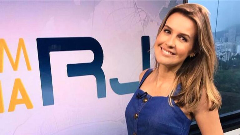 Uma mulher aparece em um estúdio de televisão, sorrindo para a câmera. Ela tem cabelo claro, na altura dos ombros, e usa um vestido azul sem mangas com botões frontais. Ao fundo, há um painel com letras grandes parcialmente visíveis e, ao lado, uma janela ou telão com vista urbana desfocada. A iluminação é profissional, típica de programas de TV, destacando seu rosto e criando um ambiente moderno e informativo.