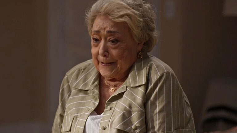 Mulher idosa de cabelos brancos curtos aparece emocionada, com expressão de preocupação, em cena interna com iluminação suave e fundo desfocado.