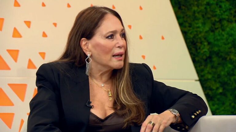 Mulher de cabelos longos castanhos, vestindo blazer preto e brincos longos, fala durante entrevista em programa de televisão, sentada em estúdio com cenário claro e elementos geométricos ao fundo.