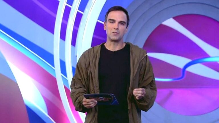Tadeu Schmidt segura um tablet e apresenta conteúdo em estúdio do BBB com cenário colorido em tons de roxo, azul e rosa ao fundo.