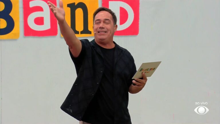 Tatola Godas sorri e acena para o público enquanto segura uma ficha em um programa de auditório. Ao fundo, um painel com letras coloridas forma parte do nome do programa Perrengue na Band, e no canto da tela aparece o selo ao vivo da emissora.