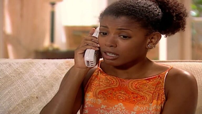 Mulher com expressão preocupada fala ao telefone enquanto está sentada em sofá dentro de sala iluminada, em cena de novela ou série