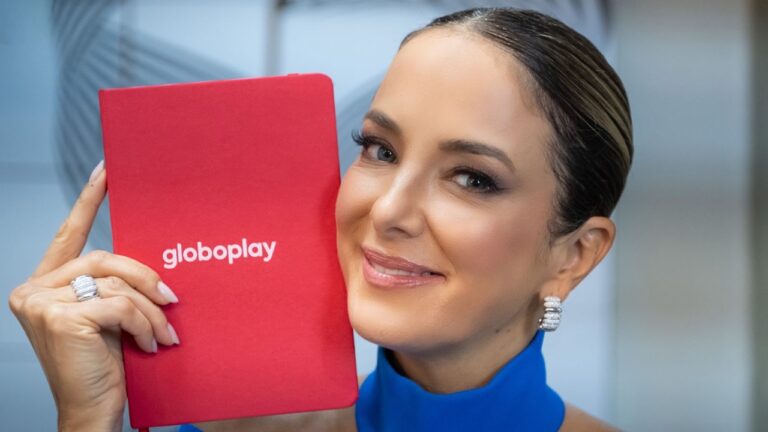 Ticiane Pinheiro, sorridente, segura um caderno vermelho com a palavra Globoplay impressa na capa, posando para a câmera. Ela usa vestido azul e brincos brilhantes, com o cabelo preso, em um ambiente interno iluminado.