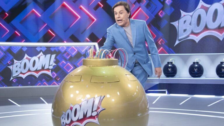 Tom Cavalcante, vestindo um terno azul, interage com um grande dispositivo dourado com fios coloridos em um estúdio de programa de auditório. Ao fundo, painéis luminosos exibem a palavra Boom! e prateleiras com objetos em formato de bomba decoram o cenário.