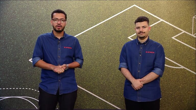 Dois apresentadores do XSports posam no estúdio com camisas azuis diante de painel com gráfico esportivo.