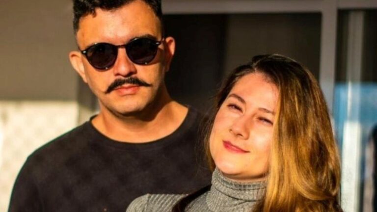 Vagner Macedo de óculos escuros e bigode posa ao lado de Bella Mantovani com suéter cinza sob luz do sol em ambiente externo