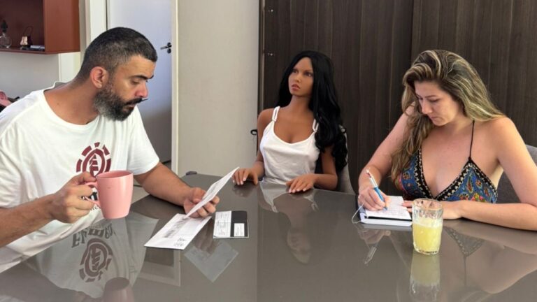 Vagner Macedo observa papel com caneca rosa, Bella Mantovani escreve em caderno e a boneca realista Kathleen senta entre eles em mesa de vidro cinza