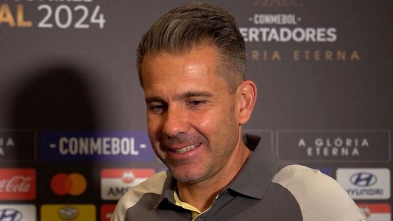 Homem de cabelo curto grisalho concede entrevista diante de painel com logotipos da CONMEBOL Libertadores, usando camisa polo em ambiente de evento esportivo.